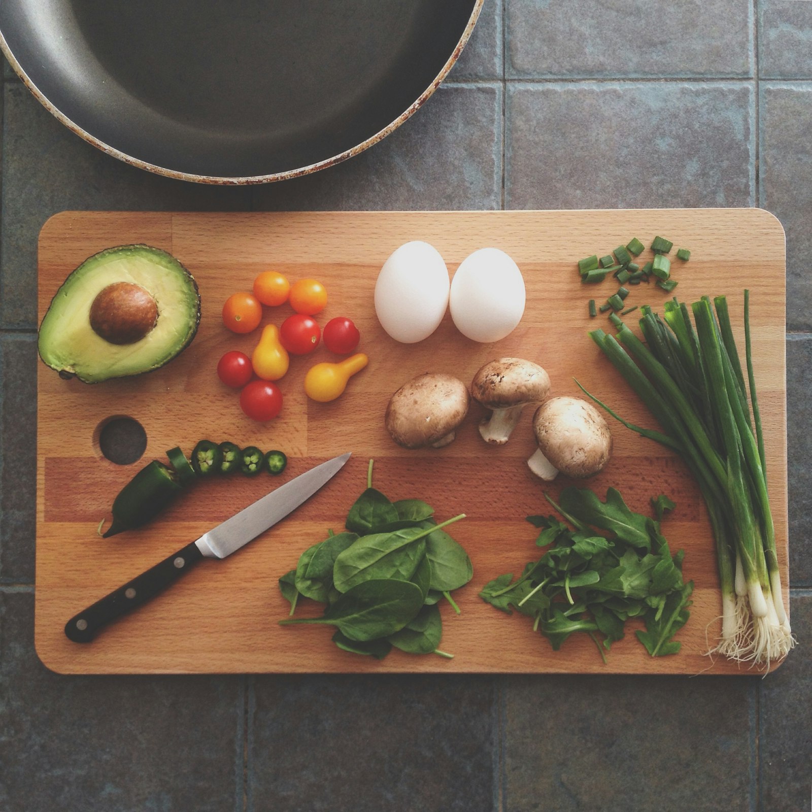 Ingredients laid out on a wooden cutting board: avocado, tomatoes, eggs, mushrooms, spinach, rocket and spring onions