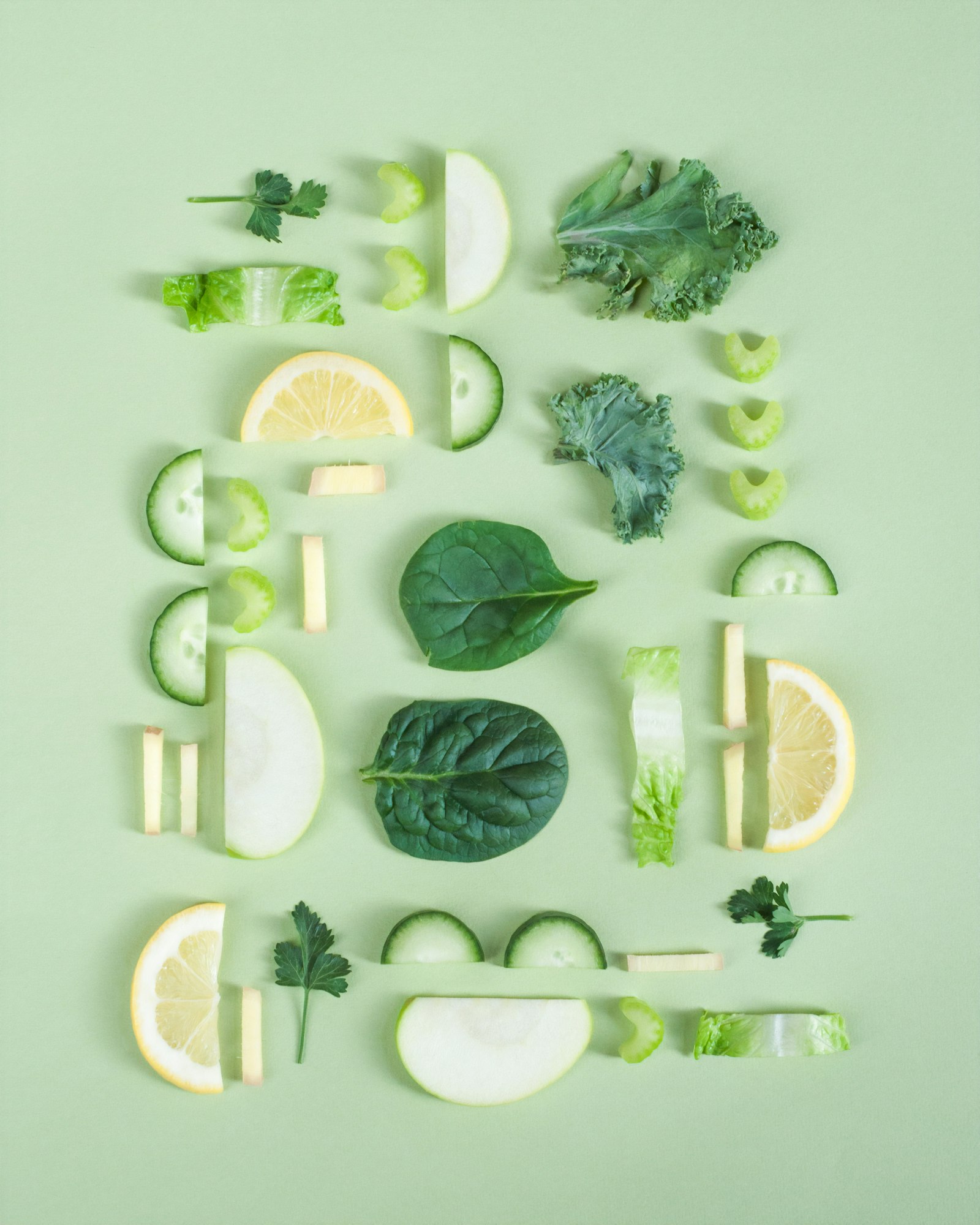 Slices of cucumber, apple, lemon, celery, kale and spinach arranged in a grid on a pale green background