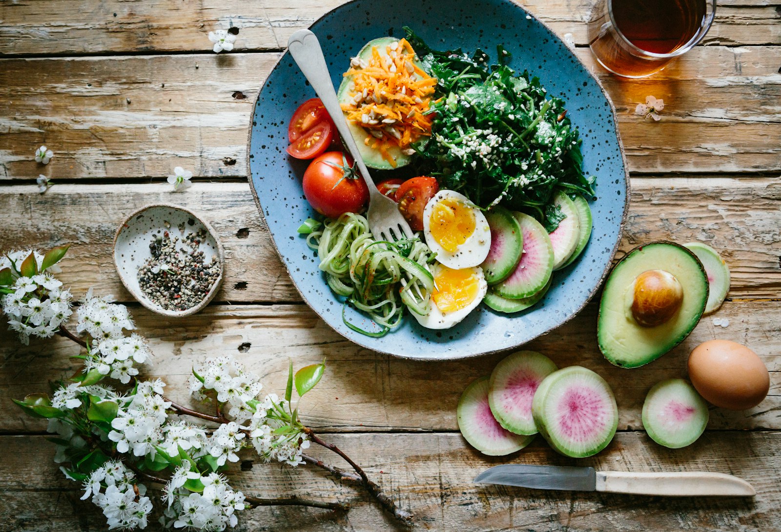 A plate of salad, soft-boiled egg, tomato and radish on a weathered wooden table, with an avocado and a knife beside it